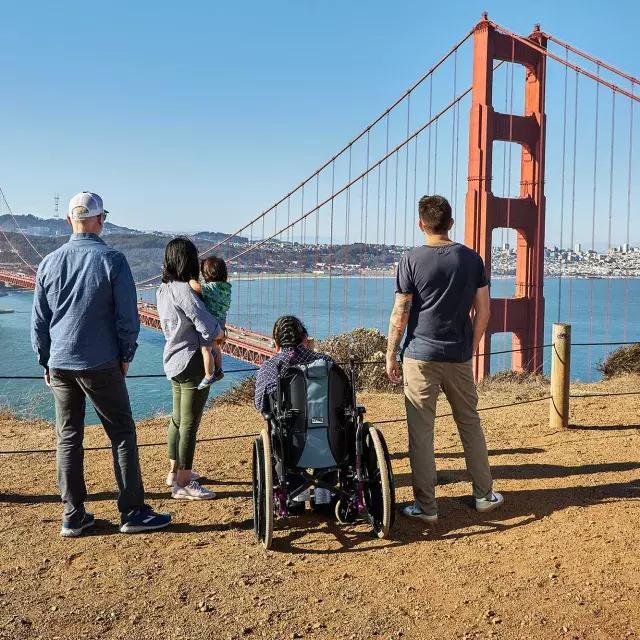 Un gruppo di persone, inclusa una persona su sedia a rotelle, è visto da dietro mentre guarda il Golden Gate Bridge da Marin Headlands.
