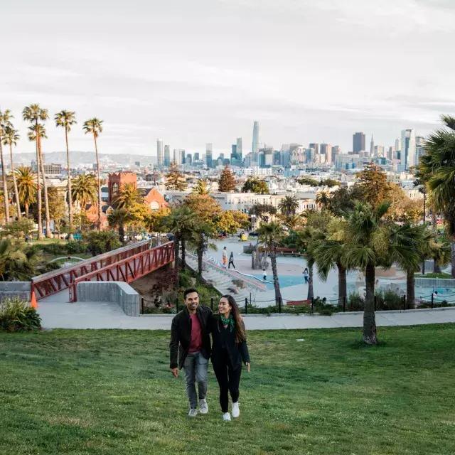 A couple walks toward the camera with Dolores Park and the 贝博体彩app Skyline behind them.